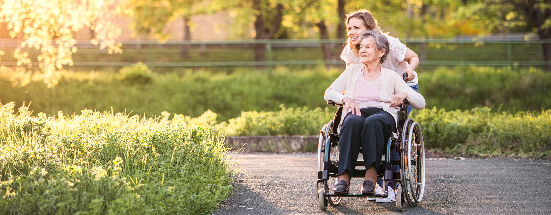 A younger woman pushes a senior woman in a wheelchair in a beautiful senior living community